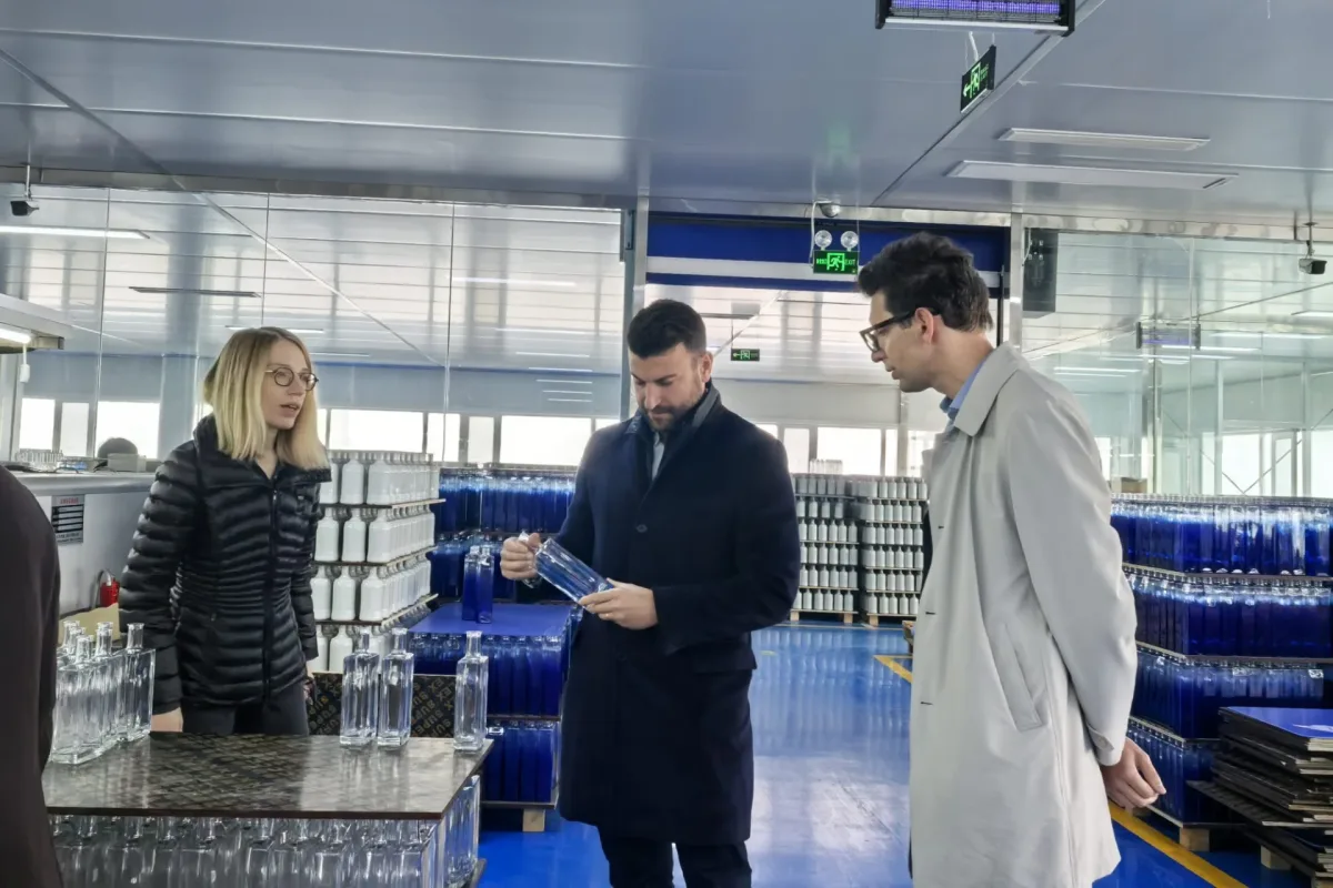 Staff inspect a clear bottle in a factory area surrounded by racks of blue and white bottles.