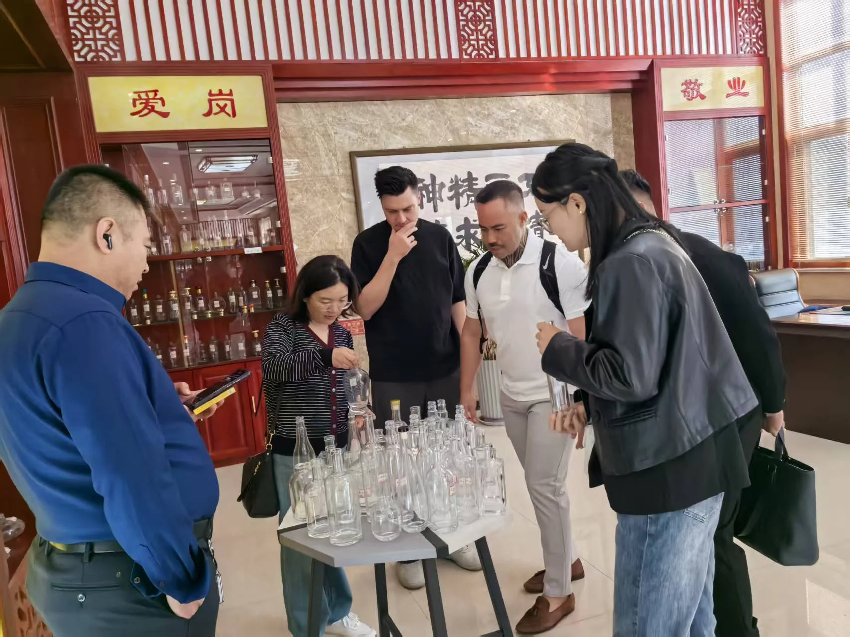 People examine a tabletop display of clear bottles in a showroom with a glass display cabinet behind them.