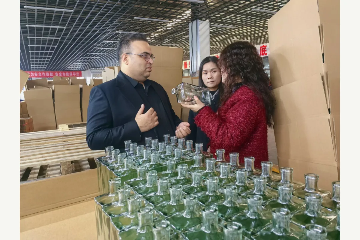 People inspect a clear glass bottle beside cartons of clear glass bottles in a warehouse.