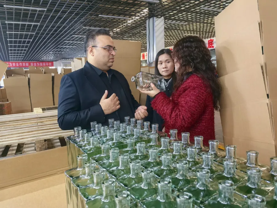 People inspect a clear glass bottle beside cartons of clear glass bottles in a warehouse.