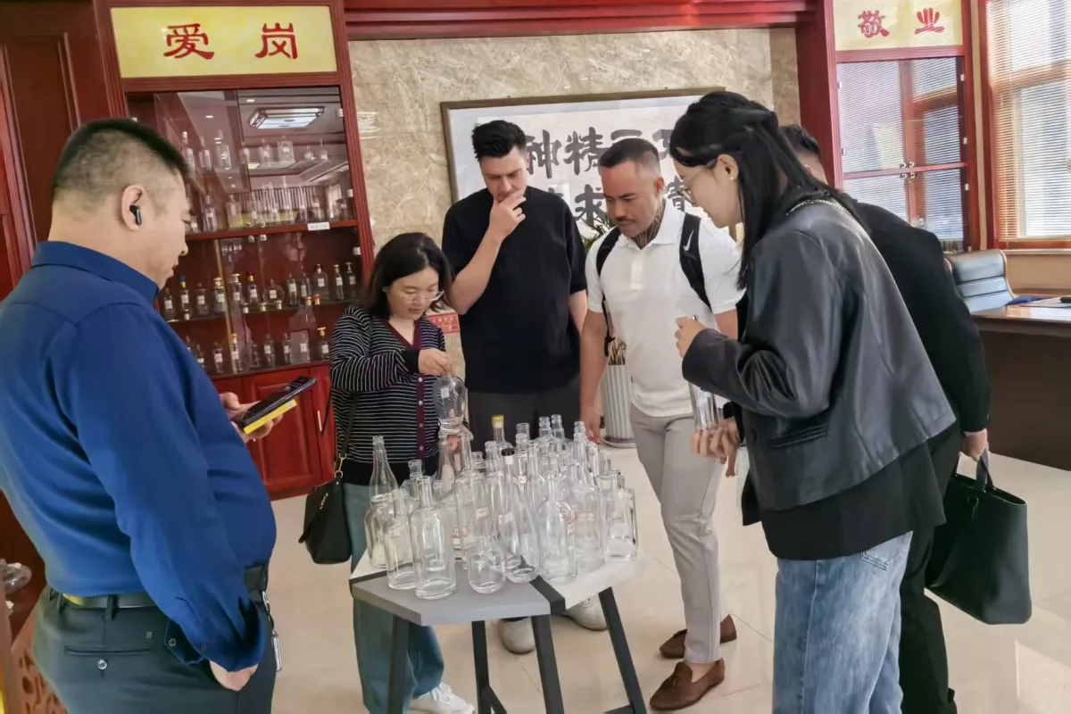 People examine assorted clear glass bottles arranged on a showroom table beside a glass display cabinet.