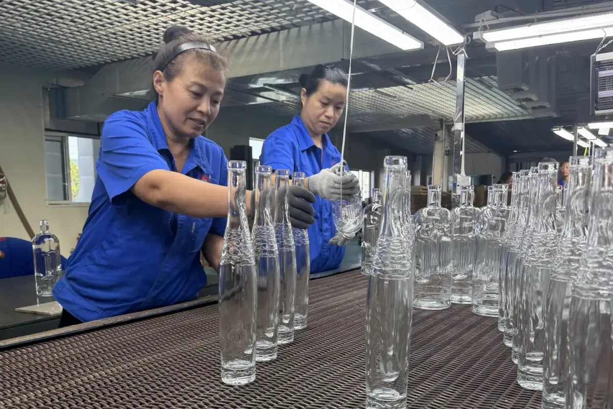 Workers inspect clear glass bottles on a factory conveyor line.