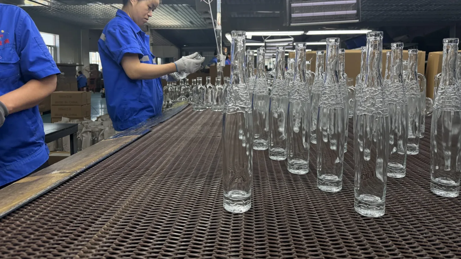 Workers inspect clear glass bottles arranged on a conveyor in a factory production area.
