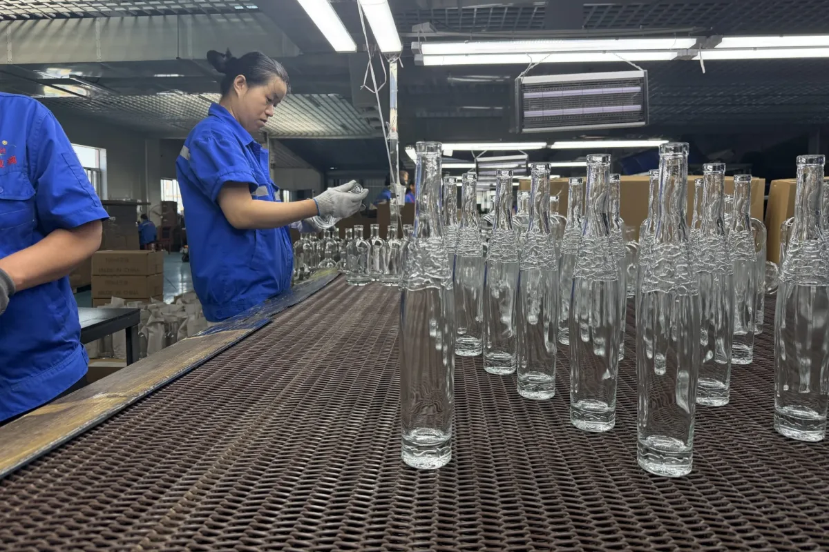 Workers inspect clear glass bottles arranged on a conveyor in a factory production area.