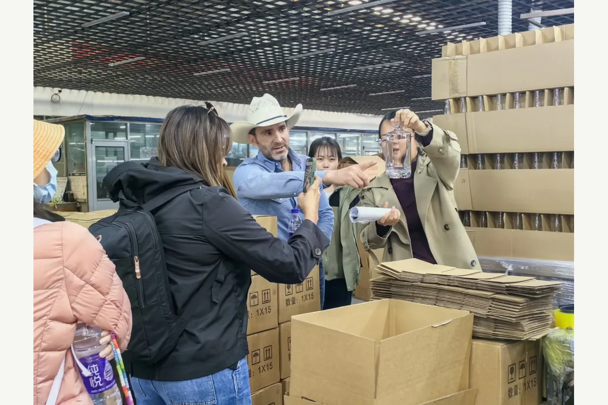 People inspect a clear plastic bottle beside stacked cartons and packaging materials in a factory area.