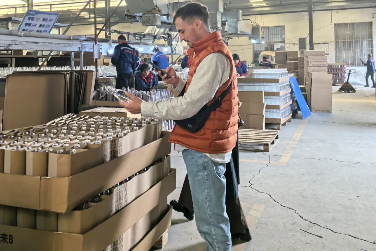 Workers inspect packaged items beside cartons and pallets inside a factory.