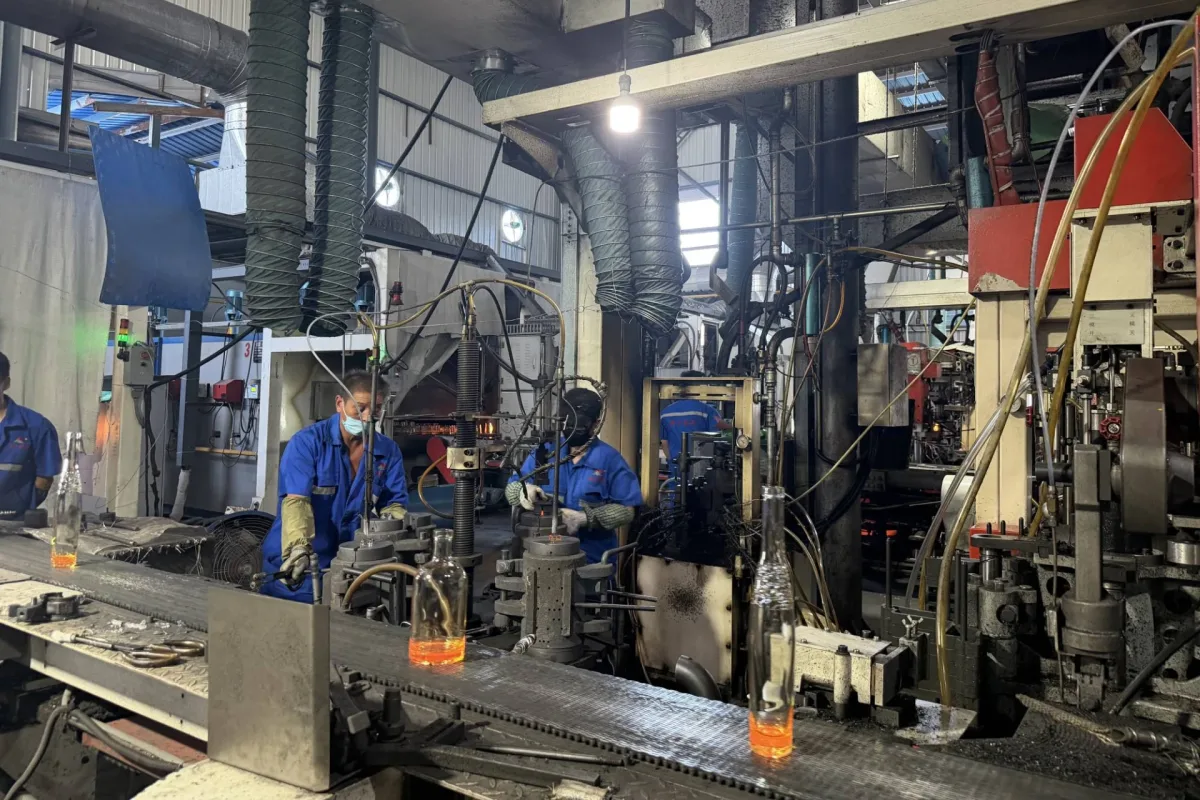 Workers operate industrial machinery as clear glass bottles move along a conveyor in a factory.