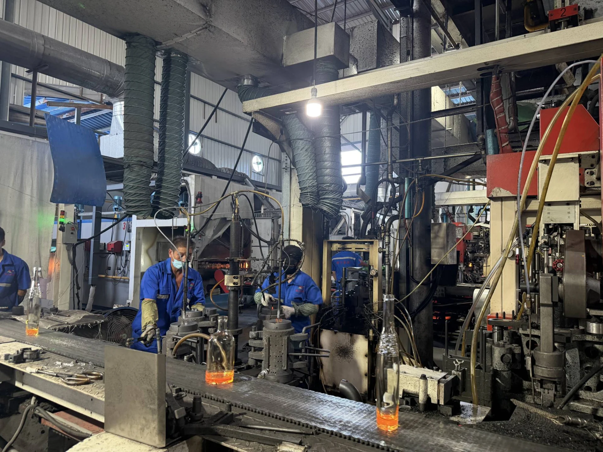 Workers operate industrial machinery as clear glass bottles move along a conveyor in a factory.
