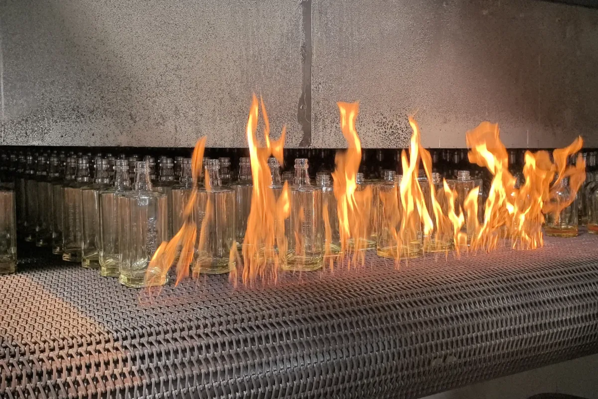Clear glass bottles moving through flames on a metal mesh conveyor inside an industrial processing area.