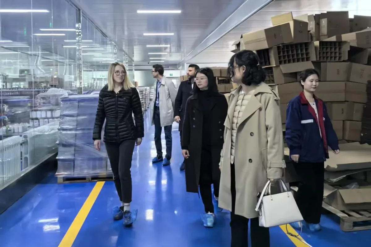 People touring a clean production facility with blue floors, yellow aisle markings, equipment behind glass, and stacked cardboard boxes.
