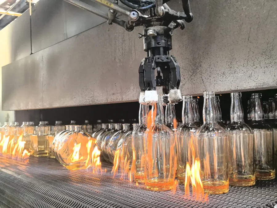 Robotic gripper handling clear glass bottles above flames on an industrial mesh conveyor.