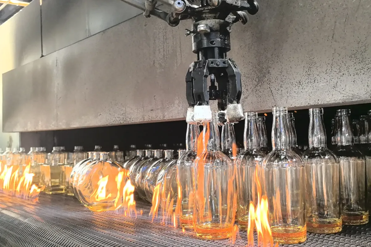 Robotic gripper handling clear glass bottles above flames on an industrial mesh conveyor.