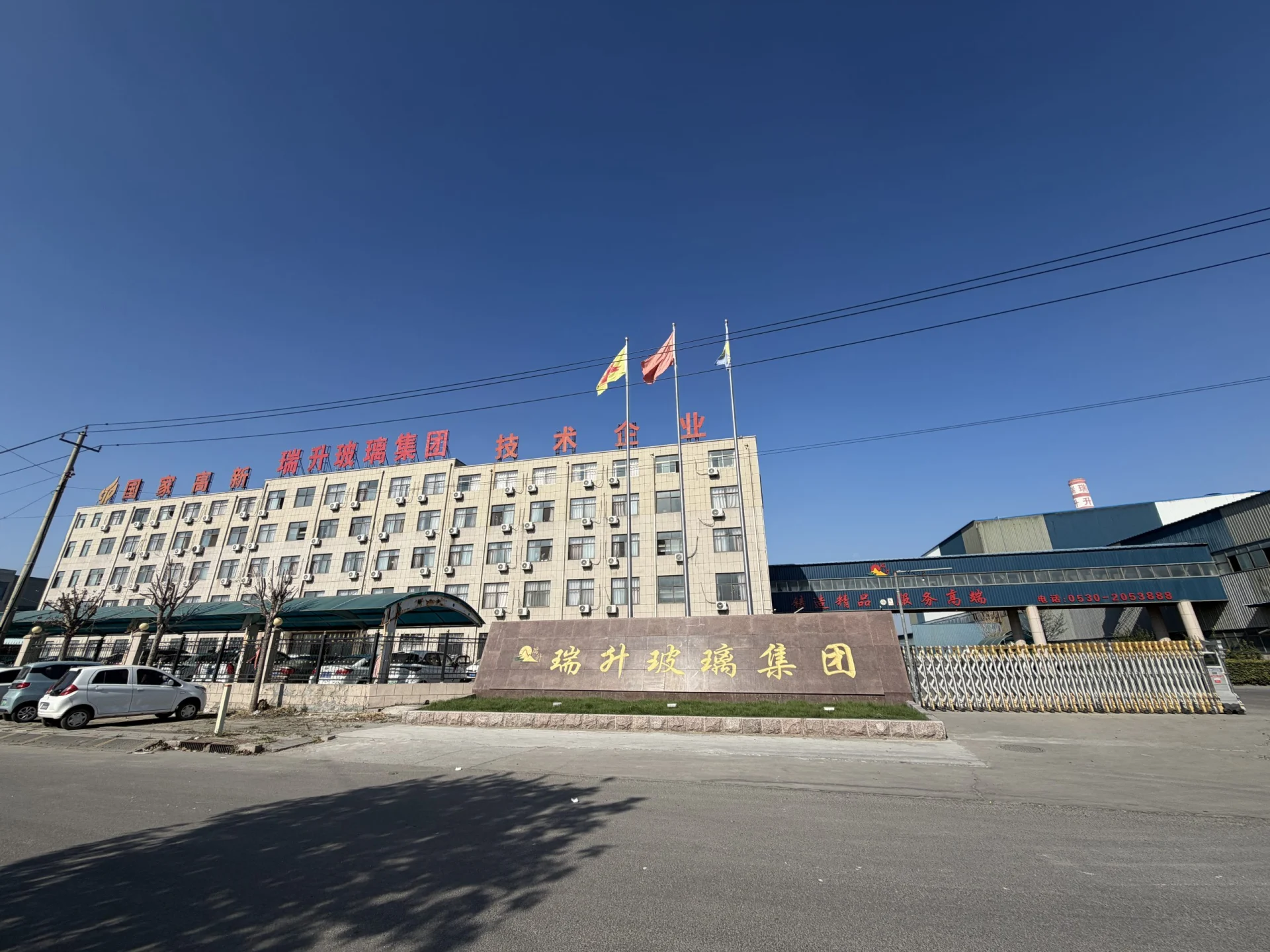 Exterior of the Ruisheng Glass Group facility with office buildings, factory structures, entrance gates, flags, and Chinese signage.