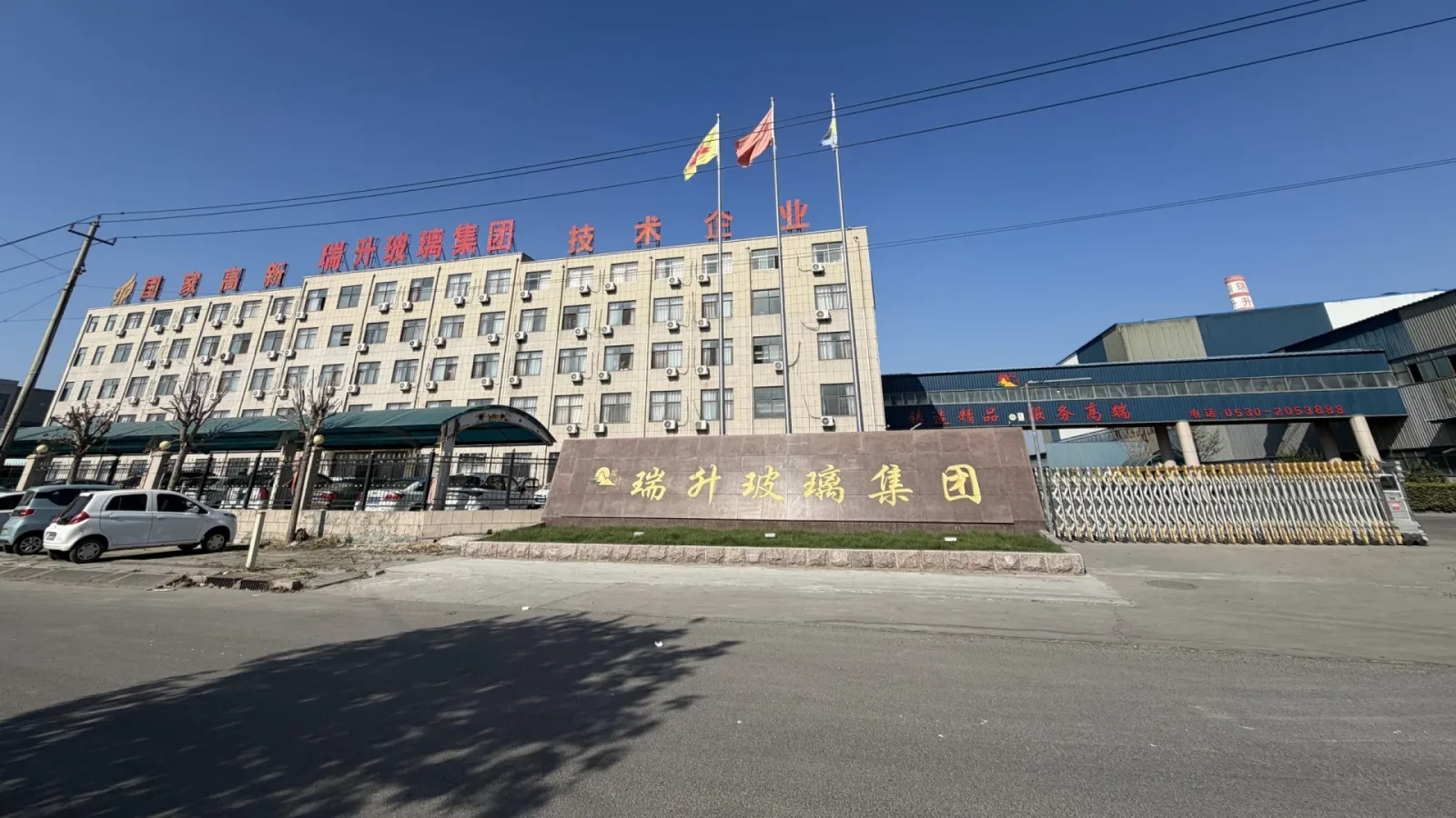 Exterior of the Ruisheng Glass Group facility with office buildings, factory structures, entrance gates, flags, and Chinese signage.