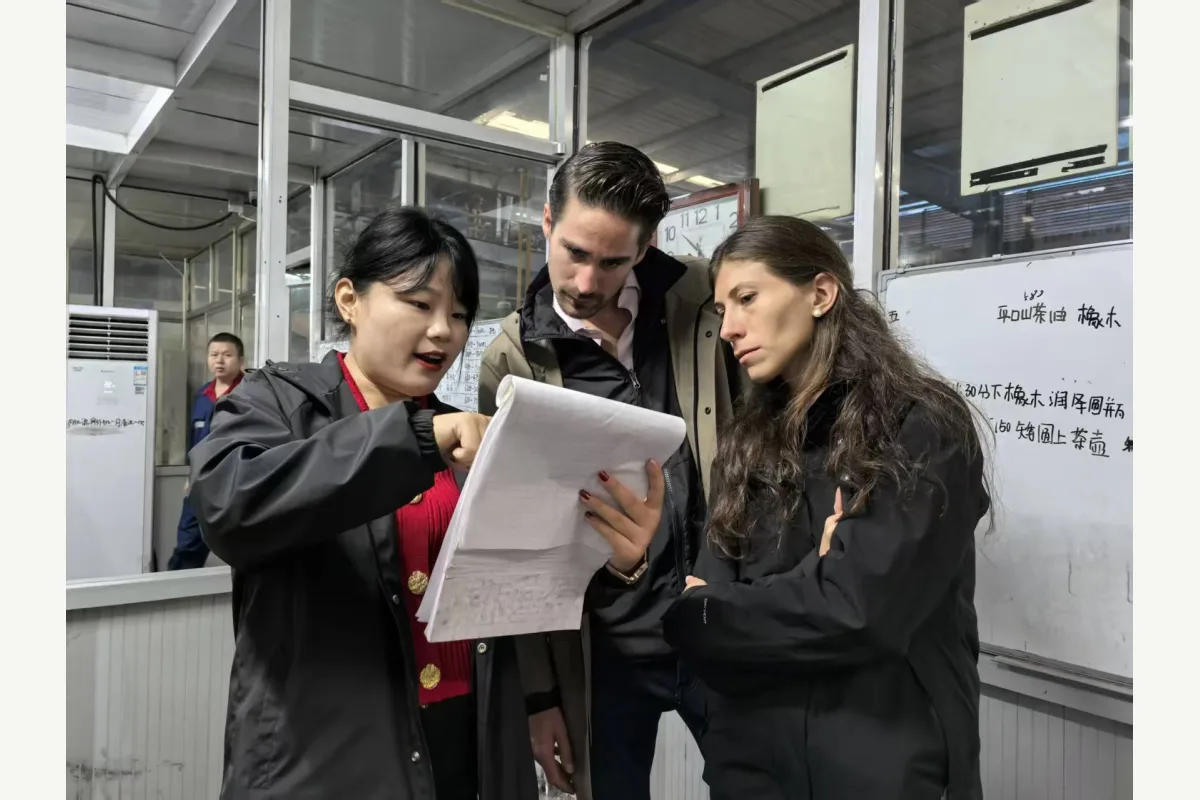 Three people review printed documents beside a handwritten whiteboard in an industrial workplace.